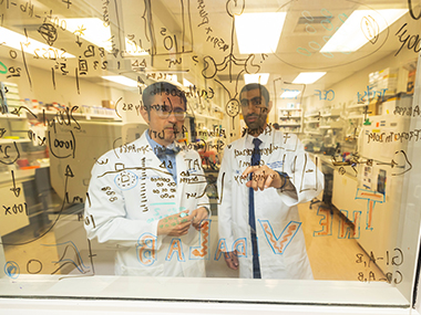 Dr, Jorge Vidal, left, discussing formulas on a transparent whiteboard with a staff member.